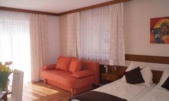 A bright room with a double bed, a black-and-white pillow, and an orange sofa. Large windows let in plenty of daylight and create a friendly atmosphere.