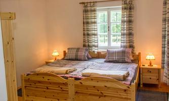 A cozy bedroom with a wooden bed and two pillows. Windows with curtains let natural light in.