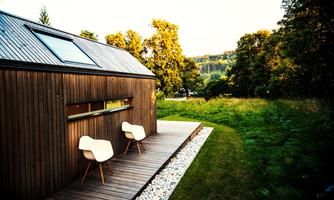 A modern wooden house with a porch and two white chairs. In the background, green trees and gentle hills can be seen.
