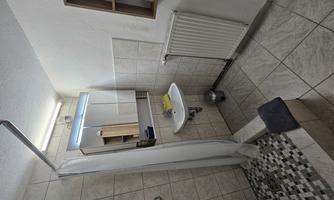 A simple bathroom with a shower, sink, and mirror. The walls are tiled, and there is a towel holder and an open shelf.