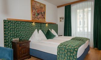 A cozy bedroom with a double bed and green decorative cushions. There is a large window front with curtains and an attractive wall mural.