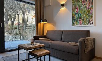 A modern living room with a gray sofa and a wooden table. Daylight is shining through the large window and the view is onto a green garden.