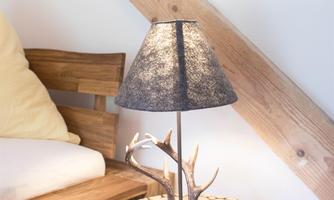 A stylish lamp with a cotton shade stands on a table made of wooden logs. In the background, a cozy sofa with a cushion is visible.
