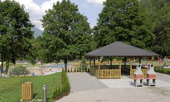 A beautiful outdoor area with trees and a pavilion. In the background, a swimming pool with bathers can be seen.