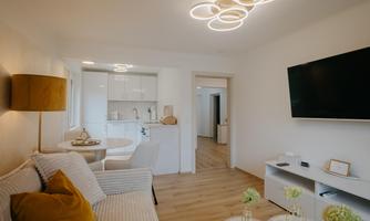 A modern living room with a cozy sofa and a large television on the wall. The open kitchen is bright and inviting, with an elegant dining table.