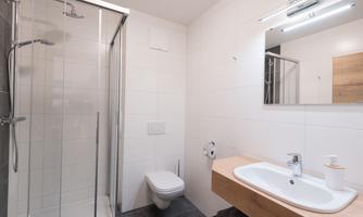 A modern bathroom with a shower, sink, and toilet. The walls are tiled in light colors, and there is a large mirror.