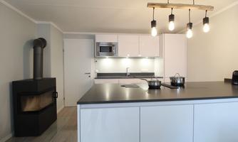 A modern kitchen with a bright, minimalist design. It features a central cooking island, a wood stove, and stylish pendant lights.
