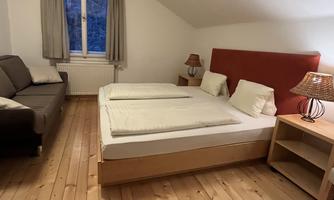 A cozy bedroom with a double bed and a sofa. The room has wood floors and ample daylight through the window.