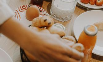 A table with various breakfast options, including pastries, fresh fruit, and beverages. A hand is reaching for the goodies in a basket.