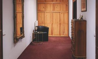 A long corridor with red carpet and neutral walls. At the end of the corridor is a wooden door and a mirror on the wall.