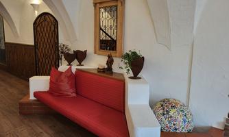 A cozy flat seating area with a red sofa and decorative cushions. In the background, there is a mirror and some plants on a wooden shelf.