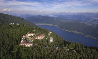 A picturesque landscape with wooded hills and a clear lake. In the background, mountain peaks and a blue sky can be seen.