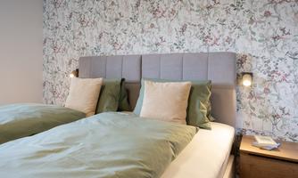 A cozy bedroom with a double bed and floral wallpaper design. The pillows and duvets in soft green tones create a relaxing atmosphere.
