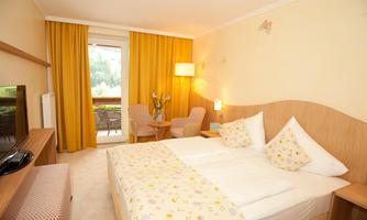 A bright room with a double bed and yellow curtains. There is a table with chairs and a view of the balcony.