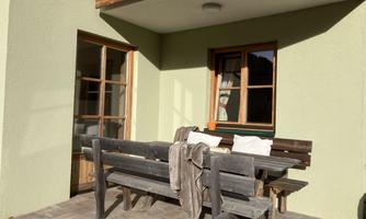 A cozy outdoor area with a wooden table group and cushions. The walls are light green, and the floor is made of paving stones.