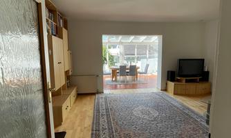 A cozy living space with a beautiful rug and light wooden flooring. In the background, there is access to the conservatory with a dining table.