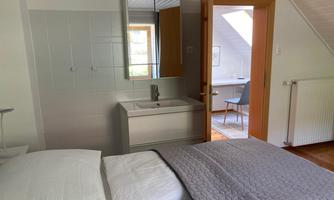 A modern bedroom with a cozy bed and a sink. Through the open door, a workspace is visible.