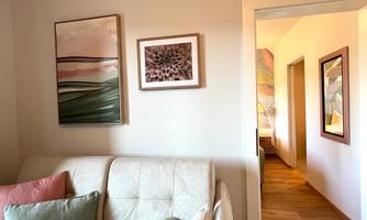 A bright and inviting living room corner with a beige sofa and decorative cushions. Two modern pictures hang on the wall, and a hallway leads to other rooms.
