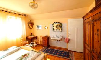 A bright bedroom with warm tones and plenty of natural light. There is a bed, a table, and an old wooden wardrobe.
