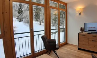 A cozy living space with large windows overlooking a snowy landscape. The wooden interior and modern furnishings create a warm atmosphere.