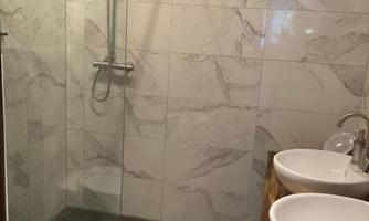 A modern bathroom with a glass shower and two sinks. The walls are designed in light marble, and the floor is dark.