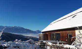 A winter landscape with a snow-covered house in the foreground. In the background, there are mountains and a clear blue sky.