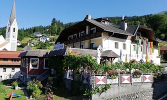 A picturesque building with colorful flowers and a balcony. In the background, you can see a church tower and a green mountain landscape.