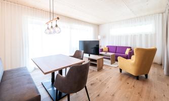 A modern living room with a dining table, a television, and comfortable seating. The large windows provide plenty of daylight and create a pleasant atmosphere.