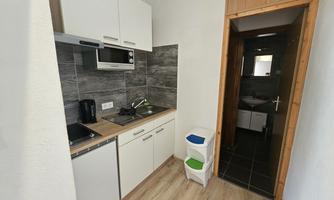 A modern kitchen with white cabinets and a gray wall. In the background, there is a hallway leading to a bathroom.