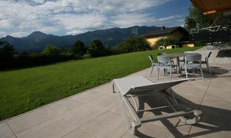 A bright terrace with a deck chair and a table with chairs. In the background, green meadows and mountains are visible under a partially cloudy sky.