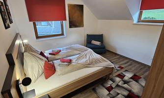A cozy bedroom with a double bed and red pillows. There is an armchair and large windows with red blinds.