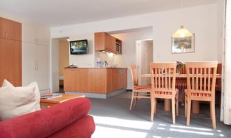 A cozy apartment with an open kitchen and a dining area. The bright furniture and the large table create an inviting atmosphere.
