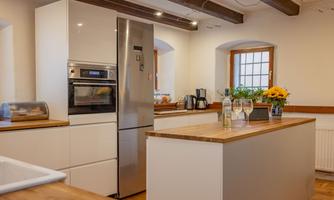 A modern kitchen with a large wooden workspace and bright walls. Daylight streams in through the windows, and wine bottles and glasses are on the countertop.