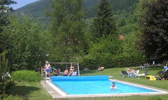A swimming pool in a green environment with mountains in the background. People are relaxing by the pool and in the sun.