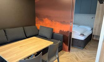 A modern room with a cozy couch and a wooden table. In the background, there is a bedroom with a bed and a stylish wall design.