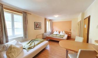 A simple guest room with two beds and a table. There are windows with curtains and a warm wooden floor.