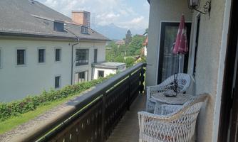 A cozy balcony with two white rattan chairs and a table. In the background, green meadows and mountains can be seen.