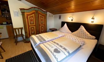 A cozy bedroom with a large double bed and traditional pillows. In the background, there is a colorful wardrobe and the walls are wood-paneled.
