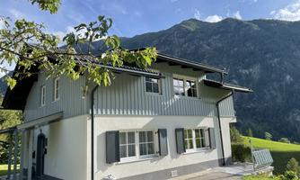 A modern house with a gray façade and white windows, surrounded by green meadows and mountains. The sky is blue with some clouds.