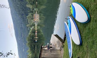 A tranquil lake with a wooden dock and stand-up paddle boards at the shore. In the background, trees and mountains can be seen.