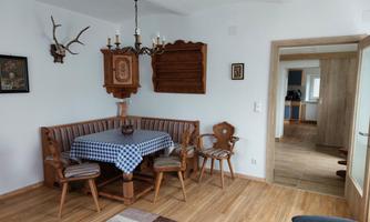 A cozy dining area with a wooden table and chairs, decorated with a checkered table runner. In the background, antique elements and an open connection to the kitchen can be seen.