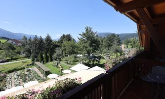 A picturesque view from the balcony of a beautiful garden with many trees and flowers. In the background, mountains and a clear blue sky can be seen.