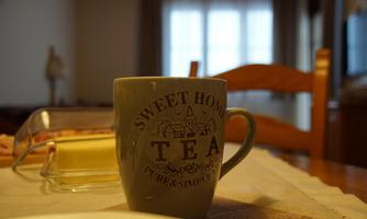 A cup with the inscription "Sweet Home Tea" is on a table. In the background, wooden chairs and a window can be seen.