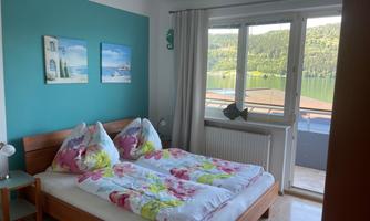 A cozy bedroom with a double bed and colorful bedding. Large windows offer a view of nature and the lake.