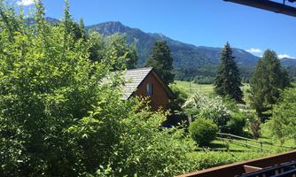 A picturesque landscape with a wooden house and lush greenery. In the background, majestic mountains rise under a clear blue sky.