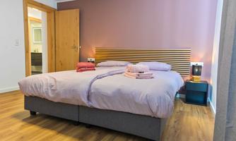 A cozy bedroom with a large double bed and modern furnishings. The walls are painted in a delicate lavender, and there are soft towels on the bed.