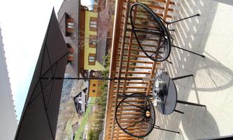 A cozy balcony with two black chairs and a table. Above the table is a sunshade, and in the background, green landscapes and houses are visible.