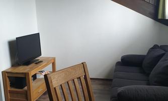 A cozy living area with a sofa and a television. The floor is wooden, and there is a dining area with a round table.