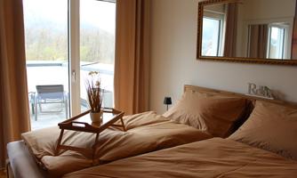 A cozy bedroom with a double bed and beige bedding. Large windows provide a view of a balcony with chairs.