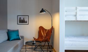 A cozy living space with a gray couch, a leather chair, and a modern floor lamp. In the background, there is a bright loft bed and a rug.